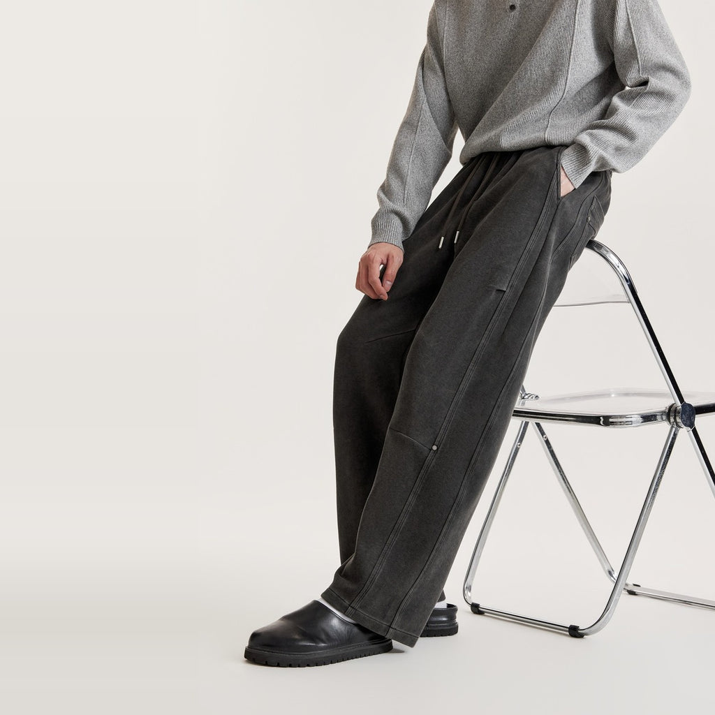 Man leaning on a clear chair in black washed wide-leg panel pants with knee seam and metal rivet detail